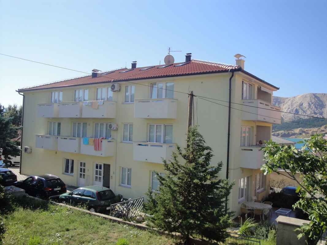 Un edificio de dos pisos con varios balcones, coches aparcados y montañas al fondo.