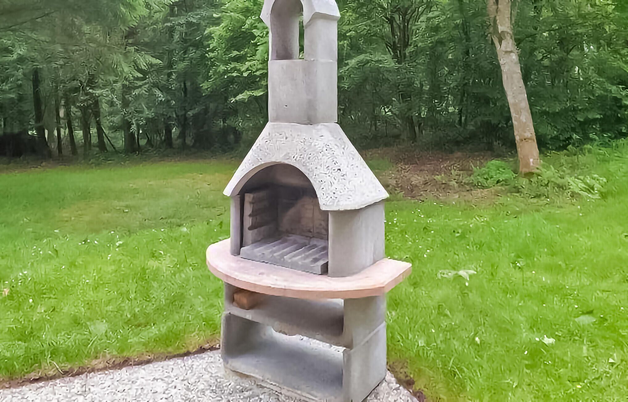 A stone-built outdoor oven with a curved front and a chimney, set in a grassy area with trees in the background.