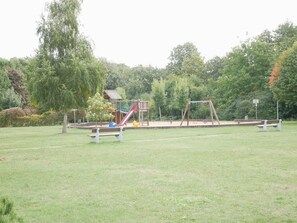 Une aire de jeux avec des balançoires et des toboggans, entourée d’arbres et d’une pelouse.