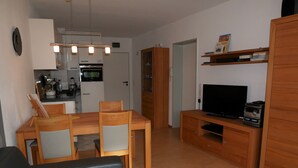 En stue med sofa, et træspisebord med stole, et fjernsyn på et træskab, og et køkkenområde med køleskab, ovn og vask.