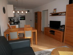 Un salon comprenant un canapé, une table à manger en bois avec des chaises, un téléviseur posé sur un meuble en bois, et un coin cuisine avec un réfrigérateur, un four et un évier.