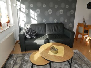 Een woonkamer met een zwarte leren bank, twee ronde houten tafels, een grijs patroon op de muur en een radiator.
