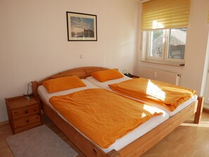 A double bed with a wooden frame, two pillows, a bedside table with a lamp, a window with blinds, and a framed picture on the wall.