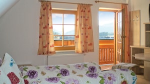Ein Schlafzimmer mit Bett, zwei Fenstern mit Vorhängen, Bergblick und einem Balkon.