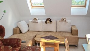 Ein gemütliches Wohnzimmer mit einem beigefarbenen Sofa, drei weichen Plüschtieren auf den Kissen, einem Holztisch und einem roten Sessel.
