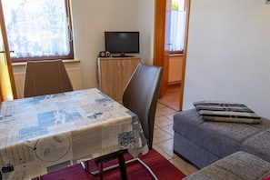 Uma sala de estar pequena com uma mesa de jantar coberta por uma toalha de mesa estampada, um sofá cinza, uma televisão sobre um armário de madeira e uma janela com vista para árvores.