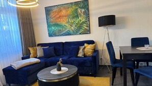 A living room with a blue sofa, a round coffee table, a painting, and a dining area with a table and chairs. - (179) 2-room apartment - Apartment house Waldhaus (Kühlungsborn)