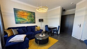 A modern living room with a blue sofa, a round coffee table, and a yellow rug. There is a dining area with a table and chairs. - (179) 2-room apartment - Apartment house Waldhaus (Kühlungsborn)