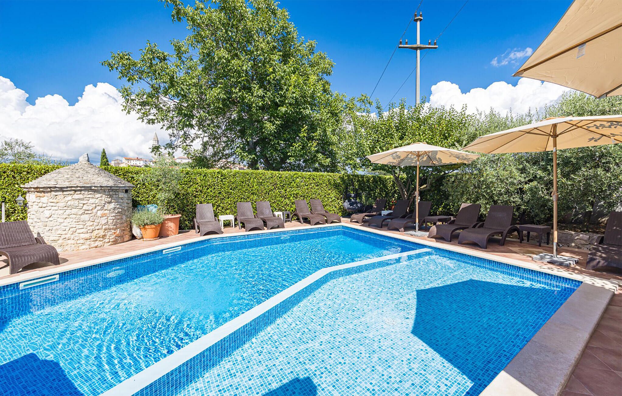 A rectangular swimming pool with clear blue water, surrounded by lounge chairs and umbrellas, set against a backdrop of trees and a stone structure.