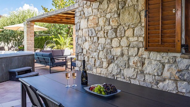 Een terras buiten met stenen muren, een houten pergola en een tafel gedekt met wijnglazen en druiven.