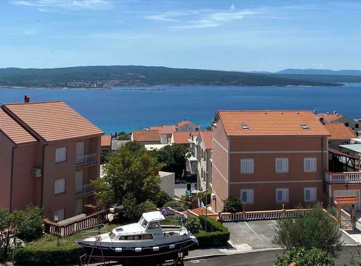 A coastal residential area with buildings, a boat docked in front, and a view of the sea and distant hills.