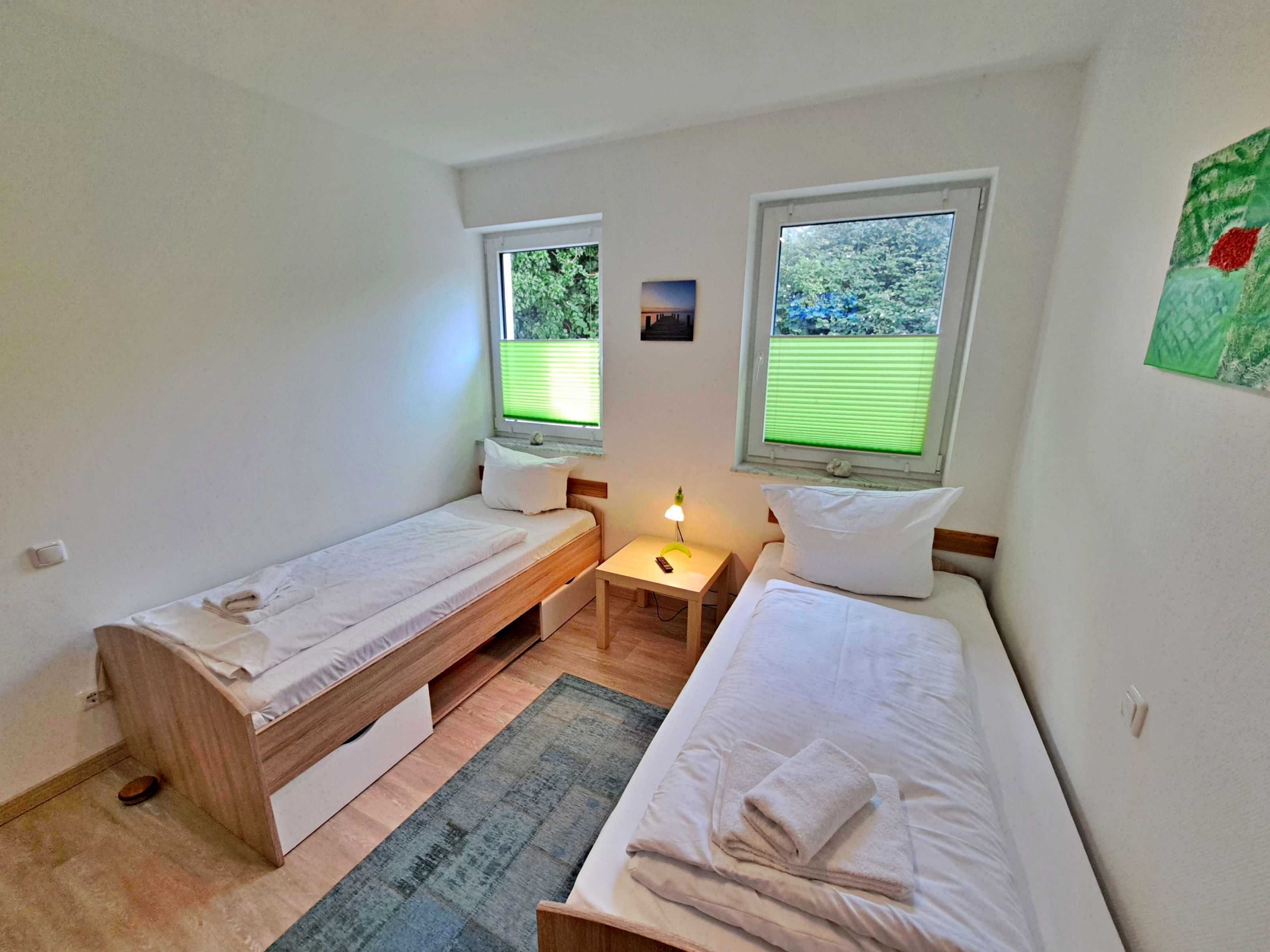 A small, clean room with two single beds, a nightstand, and a window with a view of greenery.