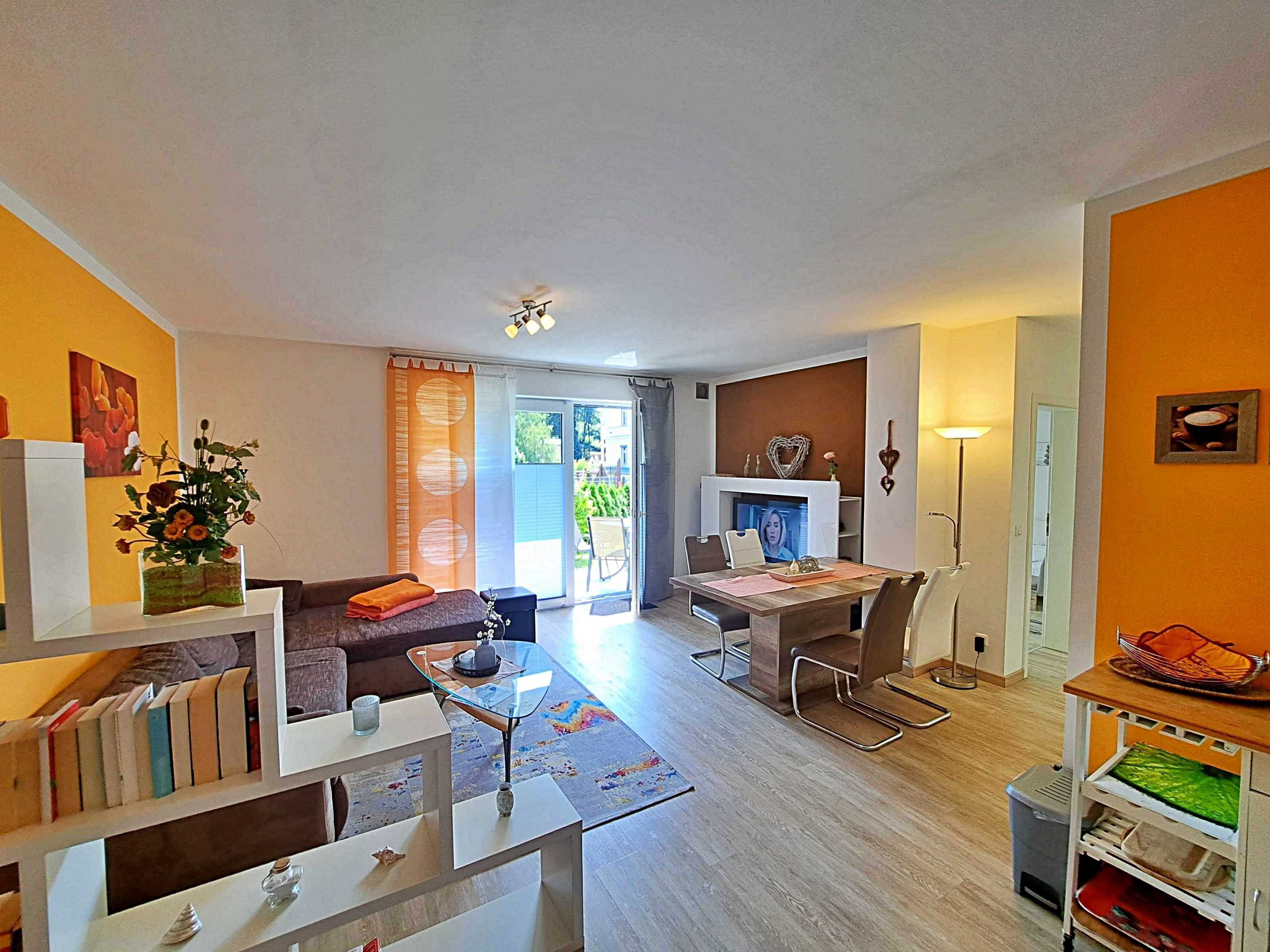 Ein modernes Wohnzimmer mit einer Couch, einem Couchtisch und einem Kamin. Es gibt einen Essbereich mit Tisch und Stühlen. Das Zimmer hat ein großes Fenster und einen Ausblick ins Freie.