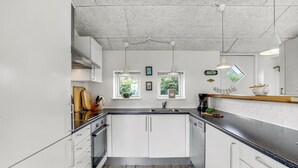 Ett modernt kök med vita skåp, en svart bänkskiva och ett golv belagt med stengolv. Det finns två fönster med växter, en hängande lampa och en kaffebryggare på bänkskivan.