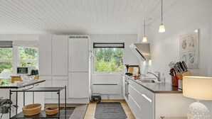 Una cocina moderna con armarios blancos, horno empotrado, microondas y pileta. Dos ventanas ofrecen luz natural y vistas a follaje exterior.