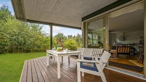 Una terraza de madera con una mesa y dos sillas, una puerta de cristal que da a una cocina, y un jardín verde exuberante.