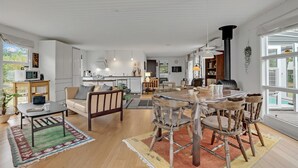 Amplia sala de estar con una gran mesa de madera para comer, múltiples sillas, un sofá y una cocina al fondo.