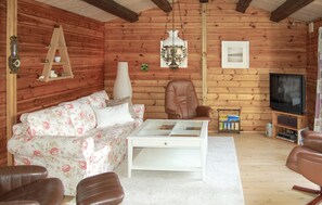 A cozy log cabin interior with a floral sofa, a leather armchair, a white coffee table, a television, and a wooden shelf with various items. - Amazing home in Rømø with kitchen (Rømø)