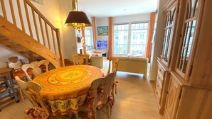 A dining area with a round table and chairs, a staircase, and a view of the street through large windows. - (269) 3- room apartment - apartment house Strandburg (Kühlungsborn)