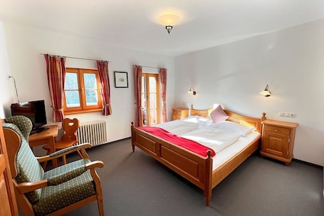 A bedroom with a wooden bed, a desk with a chair, a radiator, and two bedside lamps.. Double room Berghotel Sudelfeld - Berghotel Sudelfeld
