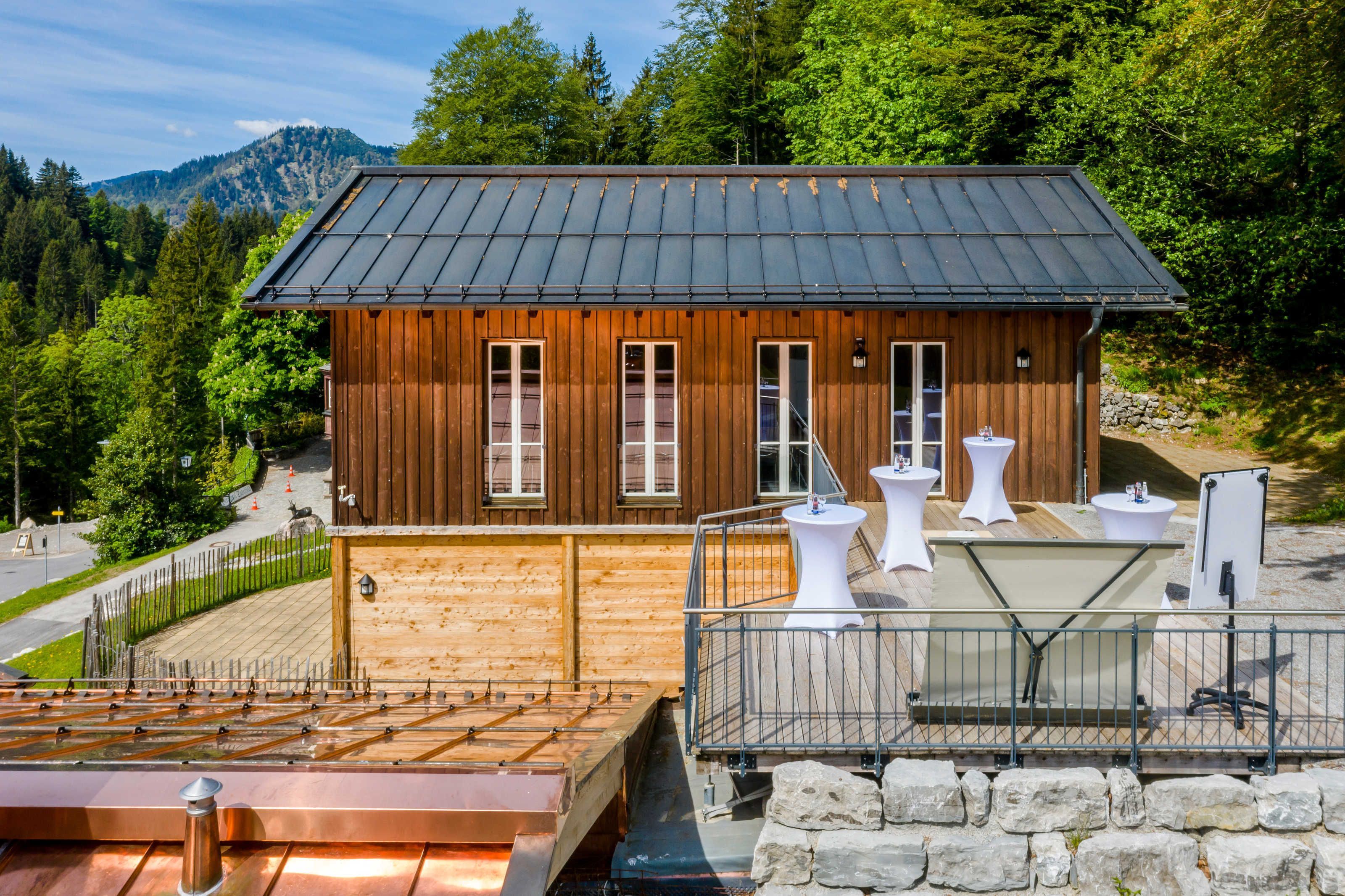 Une cabane en bois avec un toit en métal, un espace extérieur aménagé pour s’asseoir et une vue sur la forêt.