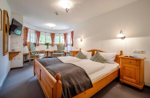 A bedroom with a wooden bed, bedside tables, a TV, and a window with curtains. - Double room Berghotel Sudelfeld - Berghotel Sudelfeld (Bayrischzell)