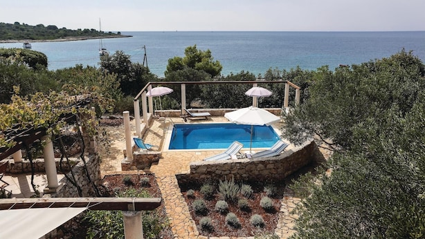 Piscina con dos sombrillas, rodeada de muros de piedra y plantas, con vista a una masa de agua y una costa lejana.
