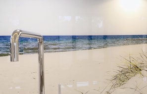 Um banheiro com torneira cromada, piso de areia e parede com cena de praia.