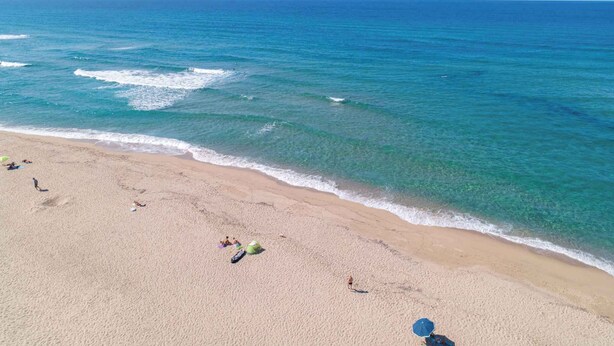 Ein Strand mit klarem türkisblauem Wasser, sandigem Ufer und einigen Personen sowie Sonnenschirmen.