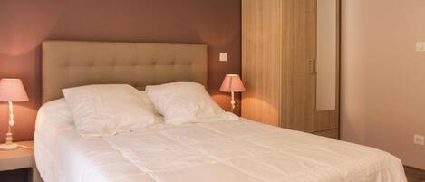 A neatly made bed with white linens, a wooden wardrobe, and two bedside lamps.