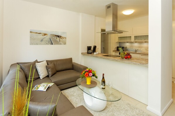 A modern living room with a brown sofa, a glass coffee table with a vase of flowers, and a magazine. A kitchen is visible in the background with a wooden countertop and various items.