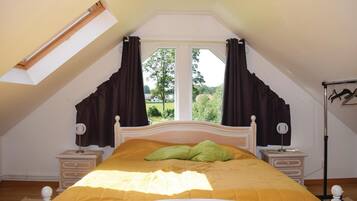 Ein Schlafzimmer mit schrägen Wänden, einem Bett mit gelber Bettdecke, zwei Nachttischen, einer Lampe und einem Fenster mit Vorhängen.