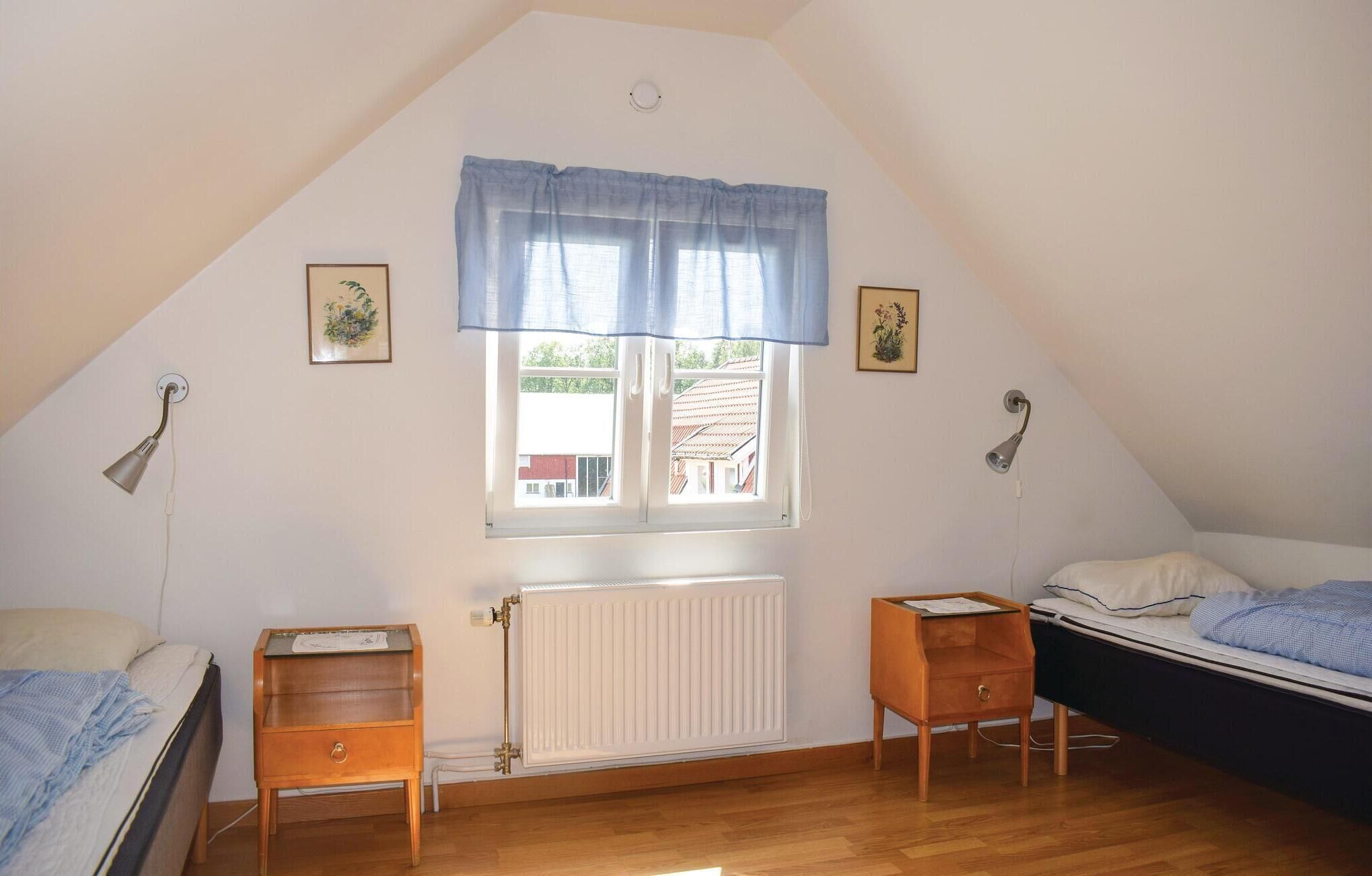 A small, well-lit attic bedroom with a sloped ceiling, a radiator, two bedside tables, a window with a blue valance, and two wall-mounted lamps.