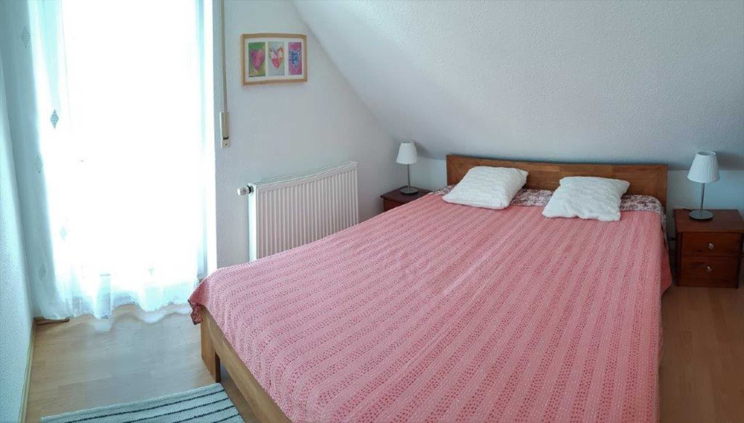 A bedroom with a wooden bed, white pillows, a pink bedspread, a nightstand with a lamp, and a radiator.