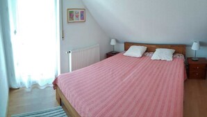 A bedroom with a wooden bed, white pillows, a pink bedspread, a nightstand with a lamp, and a radiator. - (123) 3-room apartment - Apartments Leuchtturmblick (Kühlungsborn)