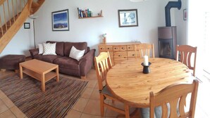 A cozy living room with a brown sofa, a wooden coffee table, a round wooden dining table with chairs, a fireplace, and a staircase. - (123) 3-room apartment - Apartments Leuchtturmblick (Kühlungsborn)