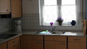 A kitchen with wooden cabinets, a window with a curtain, a coffee maker, and a sink. - (123) 3-room apartment - Apartments Leuchtturmblick (Kühlungsborn)