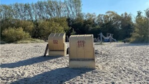 Zwei Korbstühle am Strand, mit roter Schrift auf der Rückenlehne, feiner Sand und eine Spielplatzanlage im Hintergrund.
