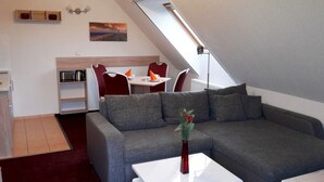 Un espace de vie moderne de style loft, comprenant un canapé d’angle, un coin repas avec des chaises rouges, un espace cuisine et une verrière.