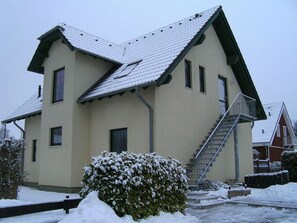 A two-story building with a sloped roof, a staircase leading to the upper floor, and a snow-covered landscape. - Rügen-Fewo 192 - Apartment (Glowe)