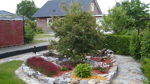 Un jardin aménagé avec une bordure en pierre, une variété de plantes et une maison en arrière-plan.