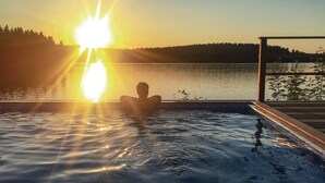 Une personne se détend dans un bain à remous, avec vue sur un lac et une forêt, au coucher du soleil.