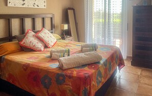 Un lit avec une couvre-lit colorée, des oreillers et une couverture. On y trouve une tête de lit en bois, une table de chevet avec une lampe et un miroir accroché au mur.