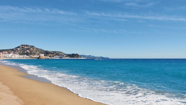 A beach with clear blue water, a sandy shore, and a hill with buildings in the background. - Awesome home in Tordera (Tordera)