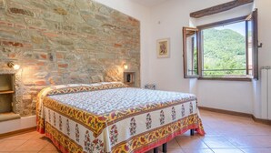 A bedroom with a stone wall, a bed with a floral bedspread, a window with a view of greenery, and a small shelf with books. - Lovely home in Dicomano FI with private swimming pool, can be inside or outside (Dicomano FI)