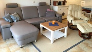 A living room with a grey sectional sofa, a white coffee table, a beige armchair, and a small white table with a lamp and speakers. - (57) 2-room apartment - apartment house Mecklenburg (Kühlungsborn)