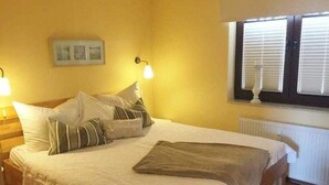 A bedroom with a bed, pillows, a bedside lamp, a window with blinds, and a radiator. - (261) 2-room apartment-Hafenstraße - Apartment holiday dreams (Kühlungsborn)
