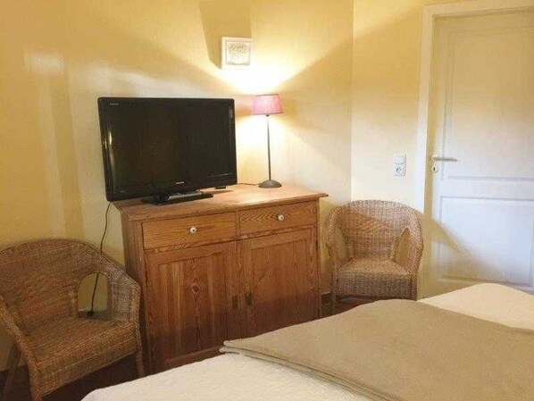 Une pièce comprenant un lit, une commode en bois avec un téléviseur, deux fauteuils en osier et une porte.