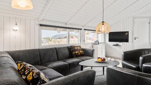 A modern living room with a sectional sofa, a round coffee table, and a black leather armchair. There is a large window with a view of the outdoors, a flat-screen TV mounted on the wall, and pendant lighting. - Cozy home in Hvide Sande with sauna (Hvide Sande)