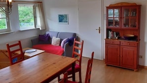 A cozy room with a wooden dining table, red chairs, a grey sofa, a red rug, a wooden cabinet with glass doors, and a window with curtains. - Apartment 1 / o. l -. Rügen-Fewo 47 (Putbus)